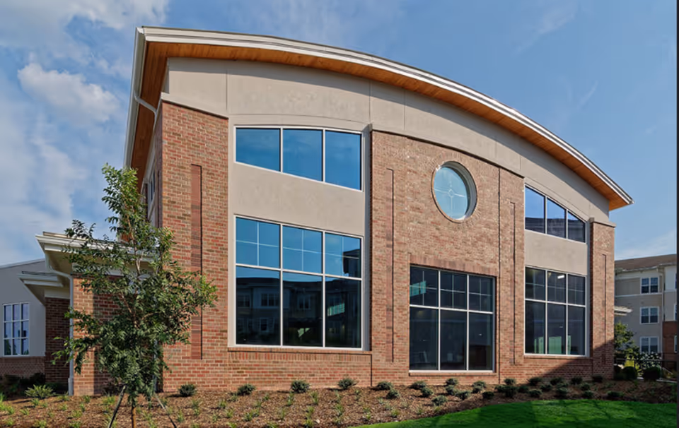 Exterior view of a modern senior living facility building with large windows, a curved roof, brick and beige walls, and landscaped greenery in front under a partly cloudy sky.