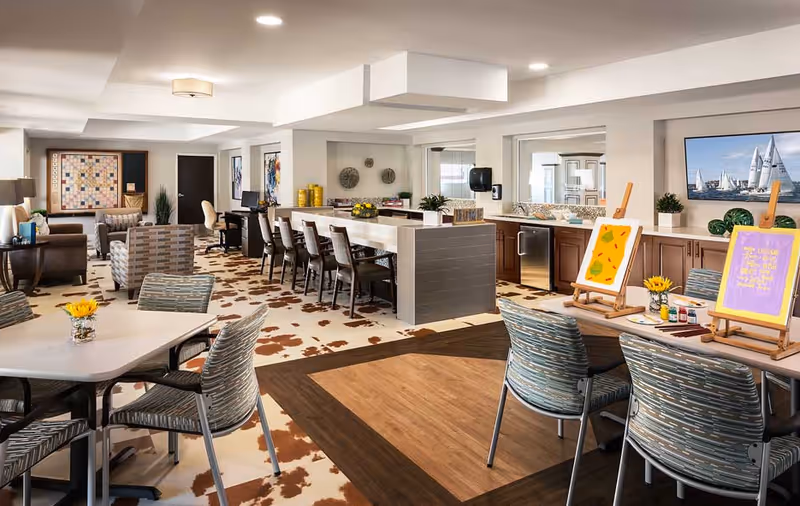 A spacious and well-lit senior living common area featuring multiple seating arrangements including tables with chairs, a long counter with bar stools, and comfortable armchairs. The room has a patterned carpet with a mix of brown and white colors, and a wooden floor section. There are art easels with paintings, decorative plants, and a large framed picture of sailboats on the wall. The area includes a kitchenette with cabinets, a dishwasher, and a sink.
