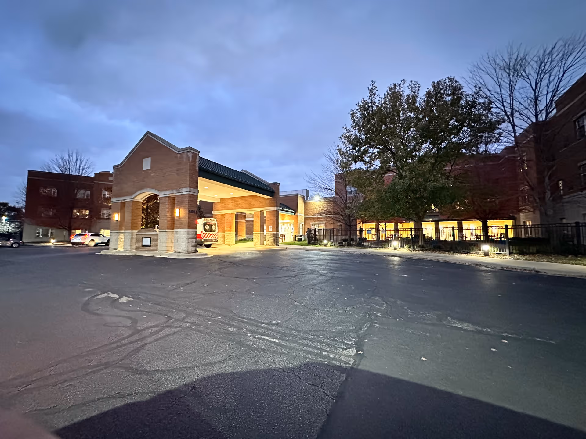 Exterior view of St. Joseph Village of Chicago at dusk, showing a brick building with a covered entrance where an ambulance is parked. The surrounding area includes a paved driveway, some trees, and lit windows.