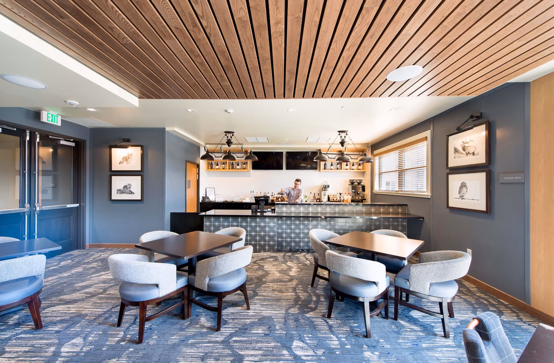 Well-lit lounge/dining area with tables and upholstered chairs facing a bar counter under a wooden slatted ceiling.