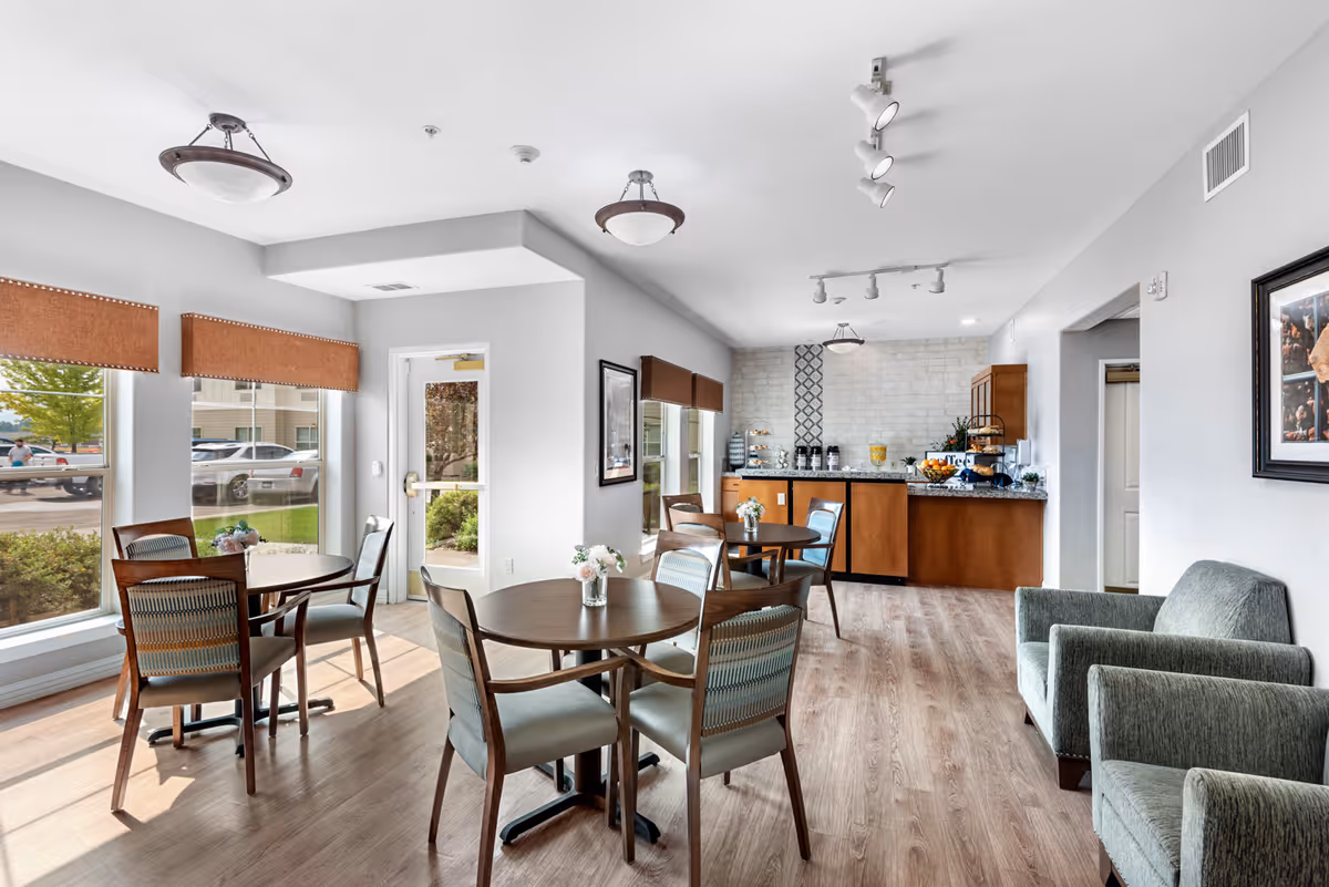 A bright and clean common area in a senior living facility featuring round wooden tables with chairs, two upholstered armchairs, large windows with brown valances letting in natural light, and a countertop area with coffee and refreshments against a white brick accent wall.