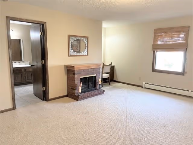 Carpeted living room with a brick fireplace topped by a mirror, a chair in the corner, a window with a blind, and an open doorway to a bathroom.
