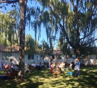 A group of elderly people sitting and standing on a grassy lawn under large trees in an outdoor area of a care facility. Some are seated on chairs arranged in a semi-circle, while others stand or walk nearby. The building of the care center is visible in the background with windows and a porch area.