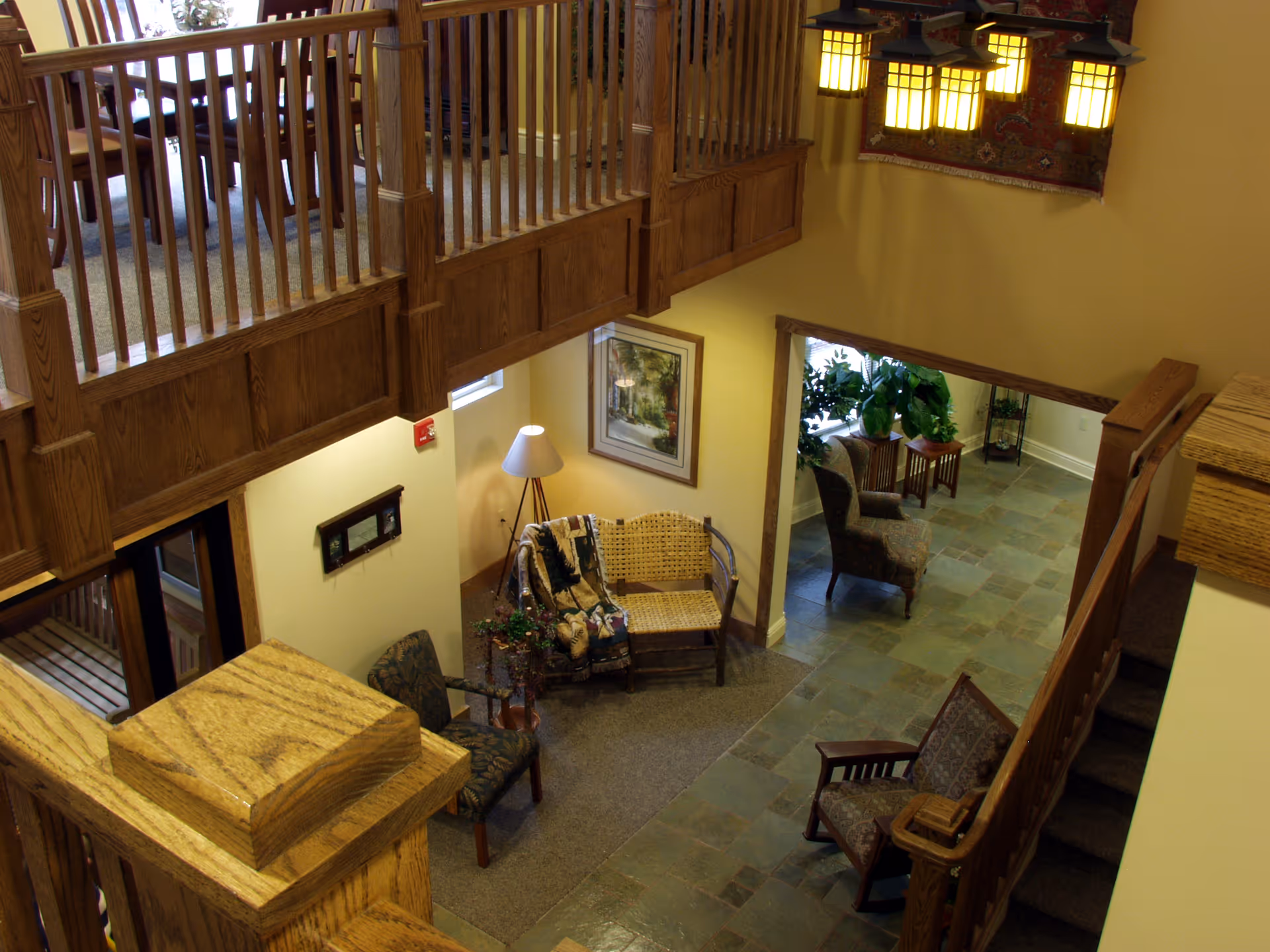 Interior view of a senior living facility showing a cozy seating area with chairs, a wicker loveseat, a floor lamp, and framed artwork on the wall. The space features wooden railings on the upper level, a tiled floor, and potted plants near a window in an adjacent room.