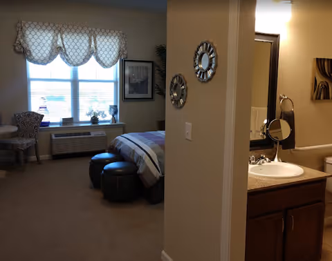 View of a senior living facility room showing a bedroom area with a bed, two round ottomans at the foot of the bed, a window with patterned valance, a chair and small table, and an air conditioning unit below the window. To the right, a bathroom sink with a mirror, towel ring, and part of a toilet is visible.