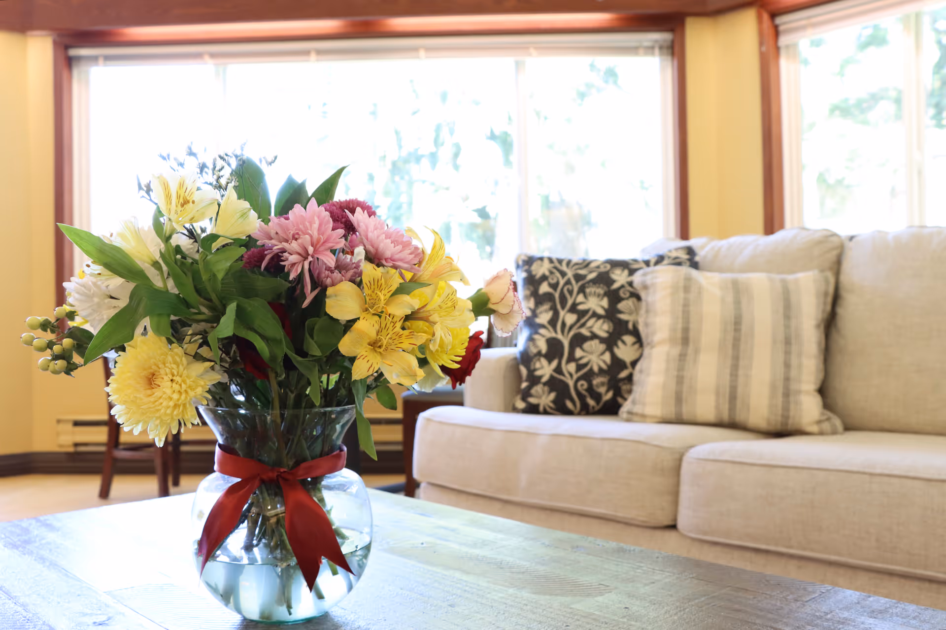 A clear glass vase with a red ribbon tied around it, filled with a colorful bouquet of flowers including yellow, pink, and white blooms, placed on a wooden coffee table in front of a beige sofa with patterned cushions in a bright living room with large windows.