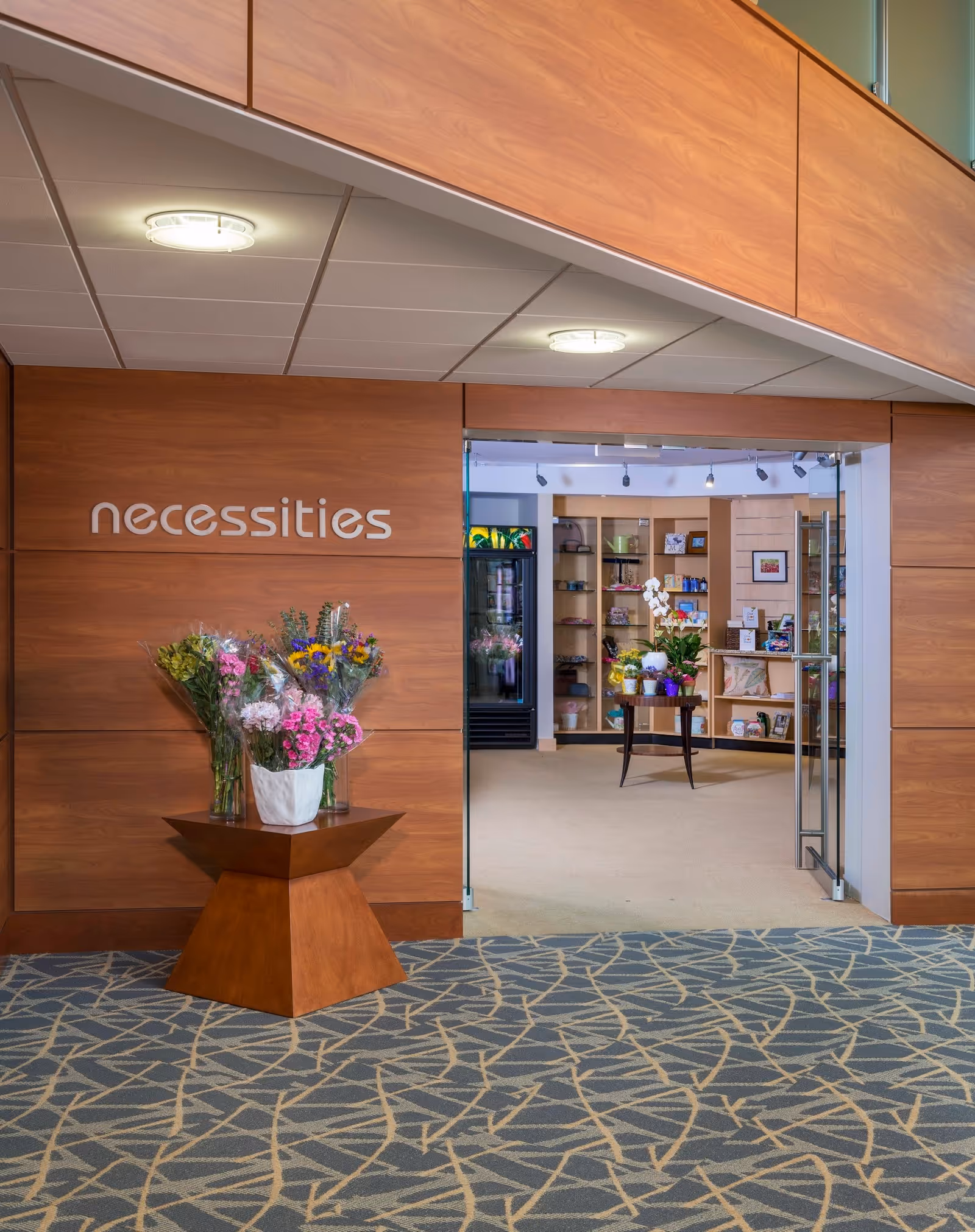 Entrance to a store named 'necessities' inside a building, featuring a wooden wall with the store name, a small wooden table with vases of colorful flowers, and glass double doors leading into the store where shelves with various items are visible.