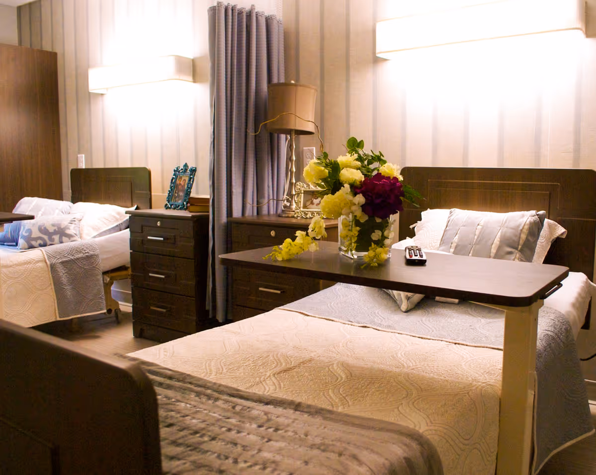 A cozy room with two hospital-style beds, each with pillows and blankets. Between the beds is a dark wooden nightstand with a lamp, a framed photo, and books. A curtain hangs in the background, and a table with a vase of colorful flowers and a remote control is positioned over one of the beds. The room has soft lighting and a warm, inviting atmosphere.