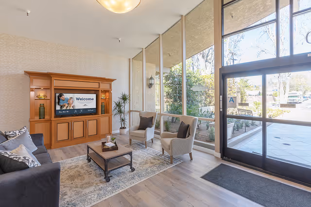 A bright and welcoming living room area with large floor-to-ceiling windows letting in natural light. The room features a dark sofa with patterned pillows, two beige armchairs, a wooden coffee table on a patterned rug, and a wooden entertainment center with a TV displaying a welcome message. Outside the windows, greenery and a bench are visible.