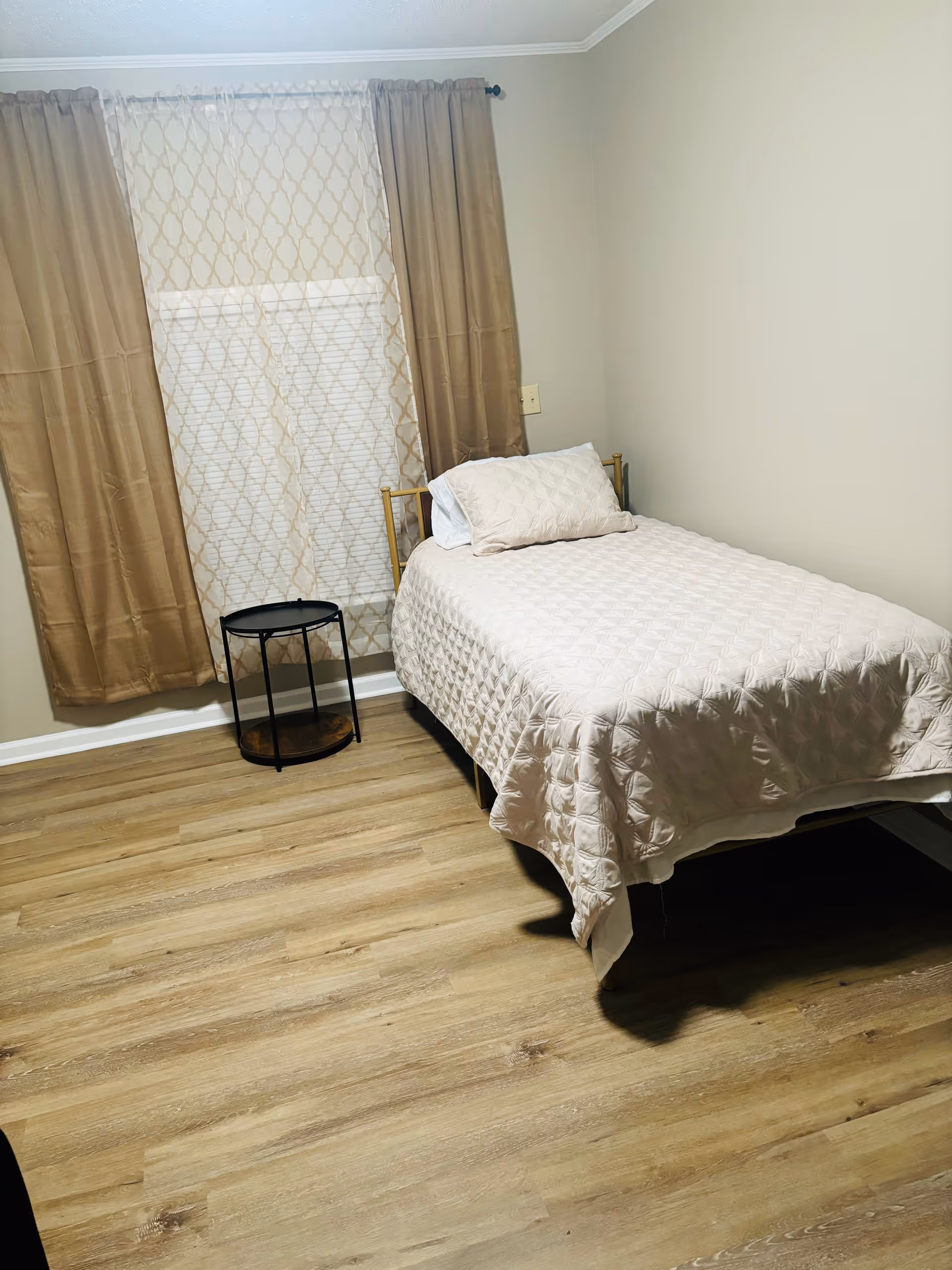 A small bedroom with a single bed covered in a light beige quilt and matching pillow. Next to the bed is a small round black side table. The room has light wood flooring and beige walls. A window with sheer patterned curtains and solid beige drapes is behind the bed.