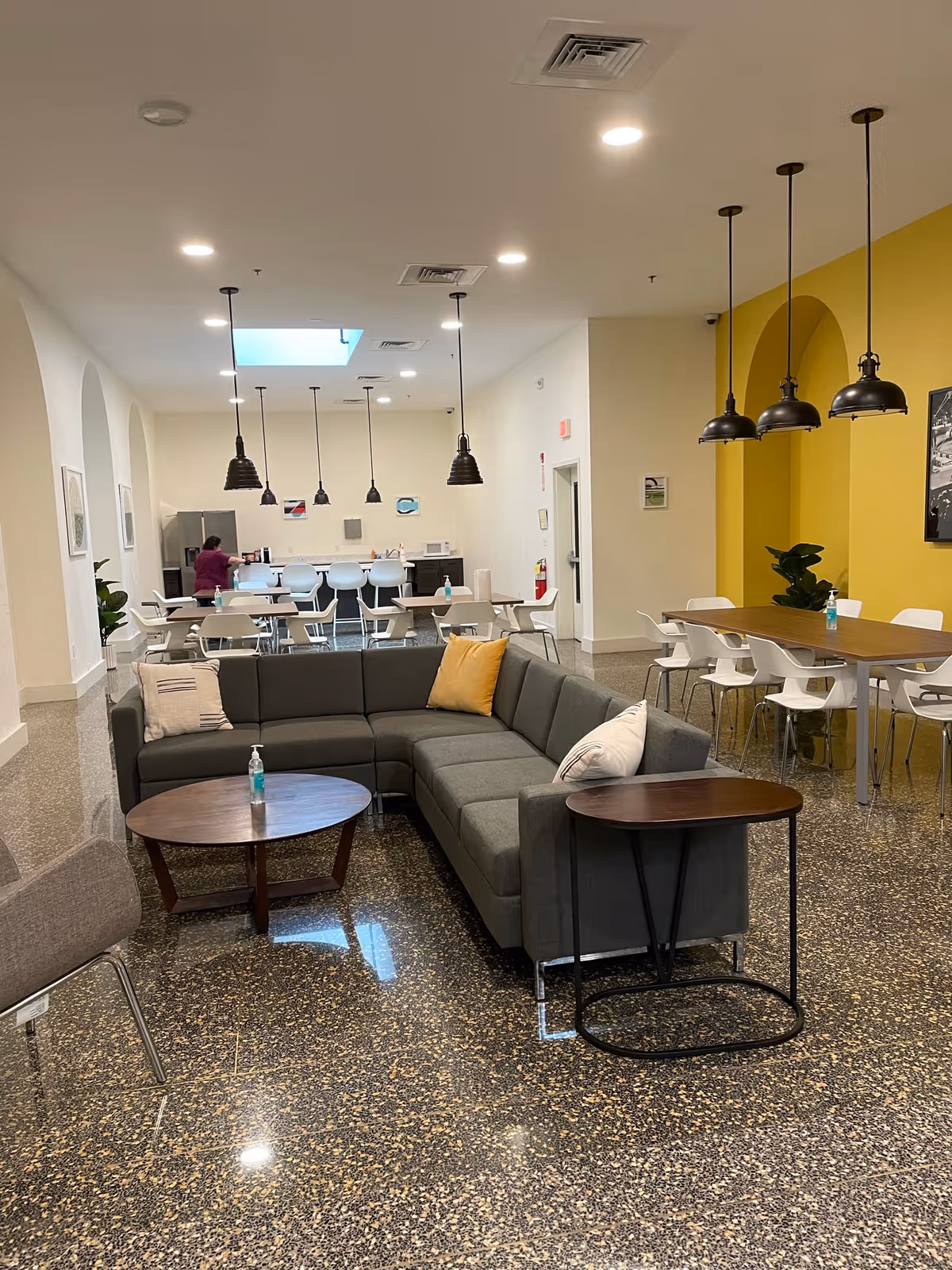 A spacious and modern common area in a senior living facility featuring a large gray sectional sofa with decorative pillows, two wooden side tables, and several white chairs around tables. The room has a polished terrazzo floor, pendant lights hanging from the ceiling, a bright yellow accent wall, and a kitchenette area in the background with a person standing near the counter.