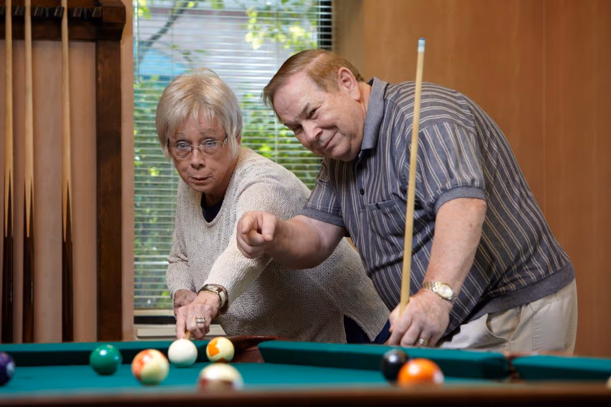 An elderly man and woman playing pool indoors. The man is holding a pool cue and smiling while the woman is aiming to strike the cue ball. They are focused on the game with a window and blinds in the background.