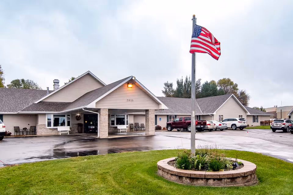 Exterior view of a single-story senior living facility building with beige siding and a covered entrance. There is a flagpole with an American flag in the foreground surrounded by a circular stone planter with greenery. Several cars are parked in the parking lot around the building under a cloudy sky.
