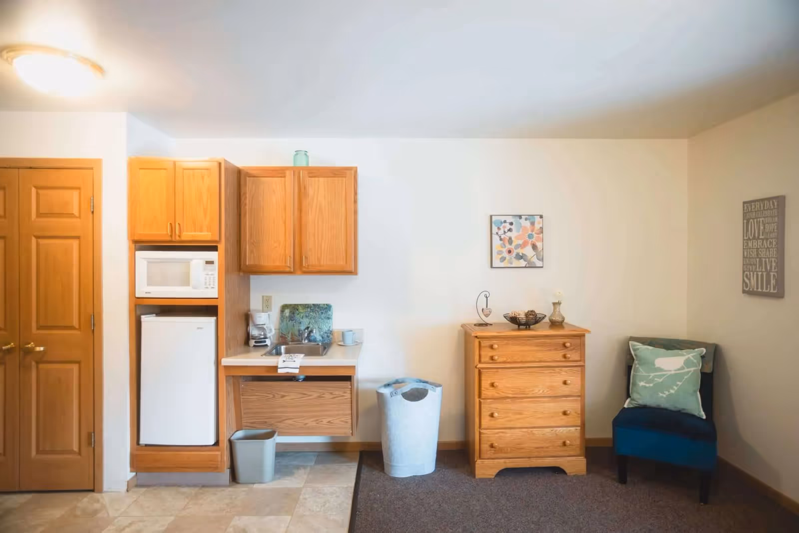 Interior view of a senior living facility room featuring a small kitchenette with wooden cabinets, a microwave, mini refrigerator, coffee maker, and a sink. Next to the kitchenette is a wooden dresser with decorative items on top, a blue upholstered chair with a bird-patterned pillow, and two wall hangings with floral and inspirational text designs.