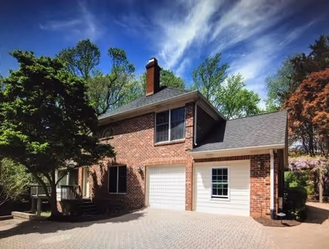 Exterior view of a two-story brick house with a chimney, a garage door, and a window. The house is surrounded by trees and greenery under a partly cloudy blue sky.