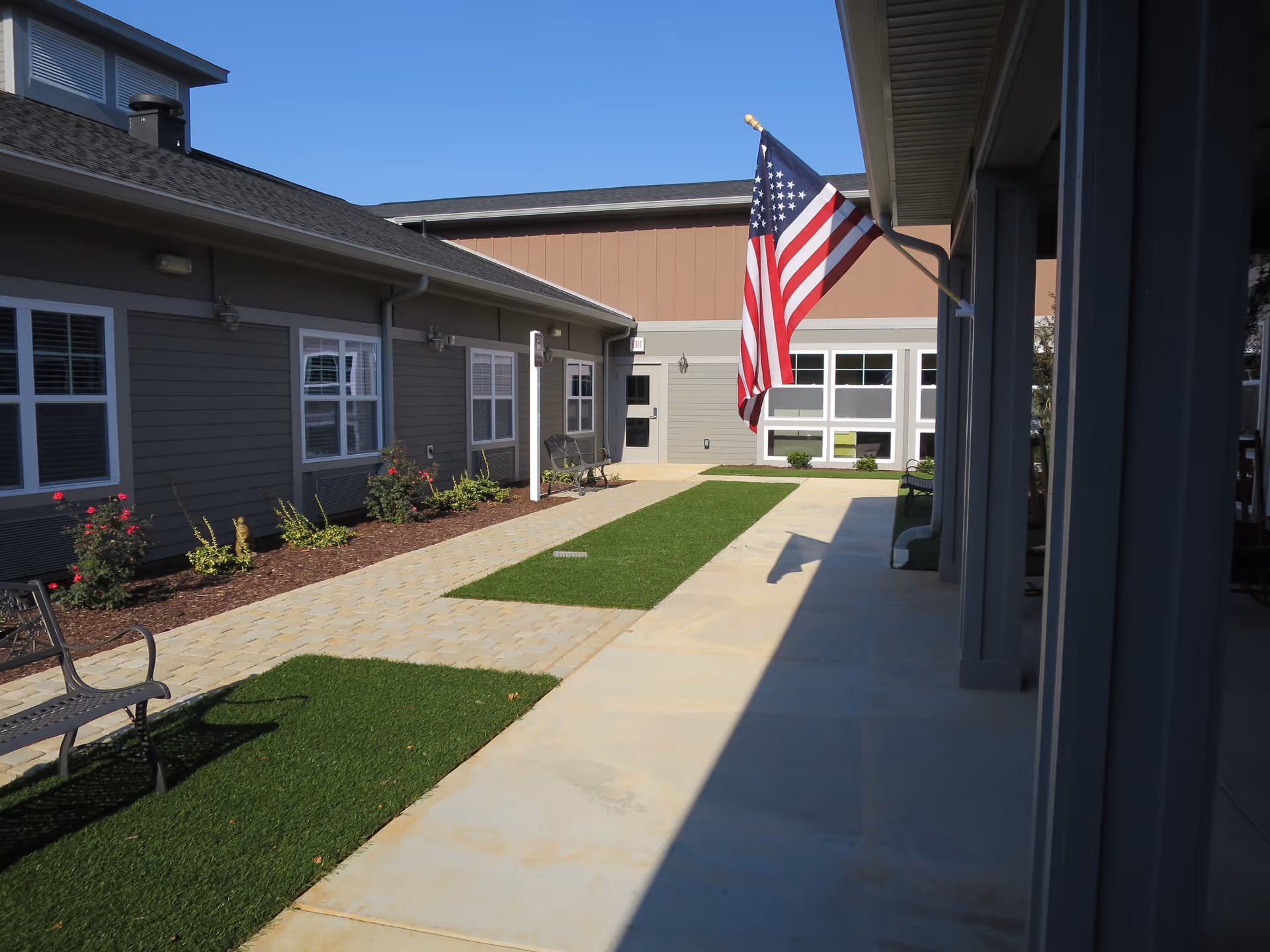 Outdoor courtyard area of a senior living facility with benches, small garden beds with flowers, an American flag mounted on the building, and a clear blue sky.