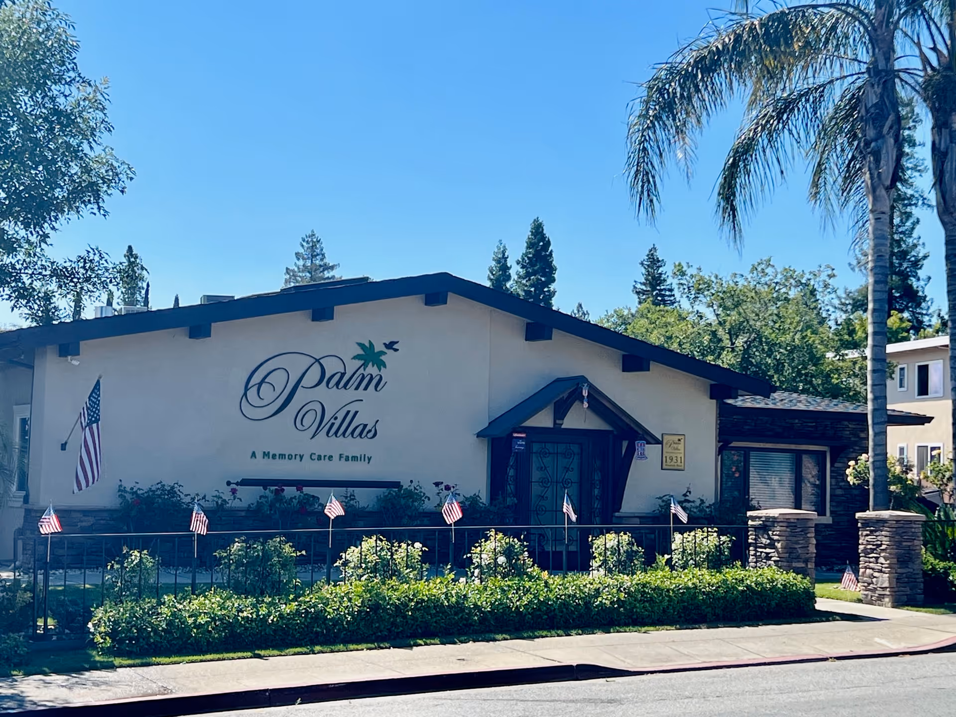 Exterior view of Palm Villas Memory Care facility building with a beige facade, dark roof trim, and a sign that reads 'Palm Villas A Memory Care Family'. The building is surrounded by greenery, small American flags, and palm trees under a clear blue sky.