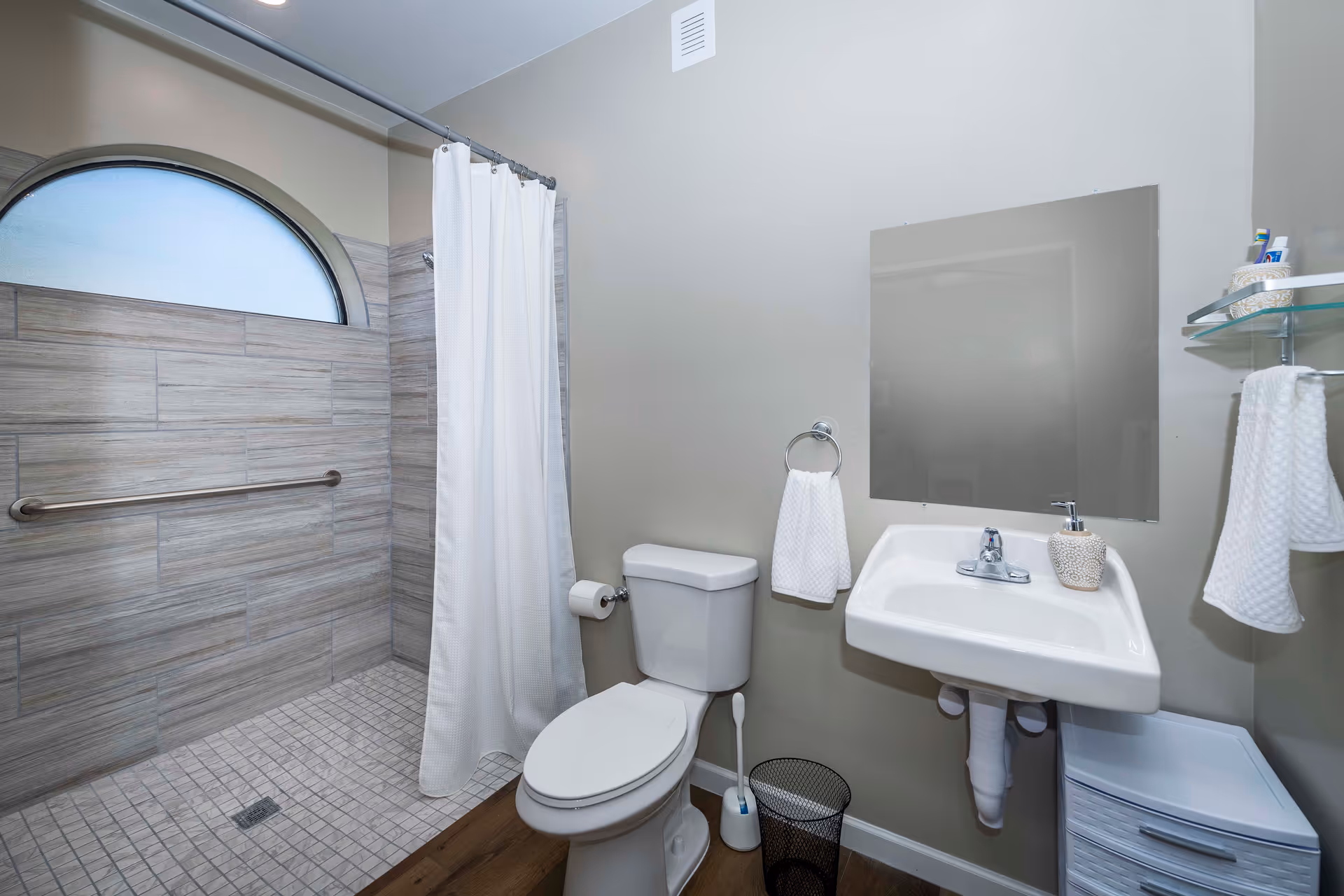 A clean and modern bathroom featuring a walk-in shower with a frosted arched window, a white shower curtain, a toilet, a wall-mounted sink with a soap dispenser, a mirror above the sink, a towel ring with a white hand towel, a small trash bin, a toilet brush, and a small cabinet with a glass shelf holding toiletries and a folded towel.