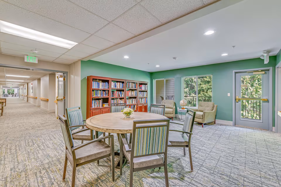 A well-lit common area in a senior living facility featuring a round wooden table with six chairs around it. Behind the table is a bookshelf filled with books against a green wall. To the right, there are two armchairs with a small side table and a globe, positioned near windows and a glass door that leads outside. The hallway extends to the left with an exit sign above a door.