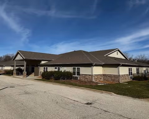 Single-story senior living facility exterior with entrance canopy, manicured shrubs, and a paved driveway.