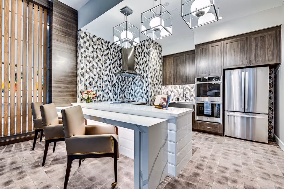 A modern kitchen with a marble island, pendant lights, stainless steel appliances, patterned tile backsplash, and chairs at the counter.