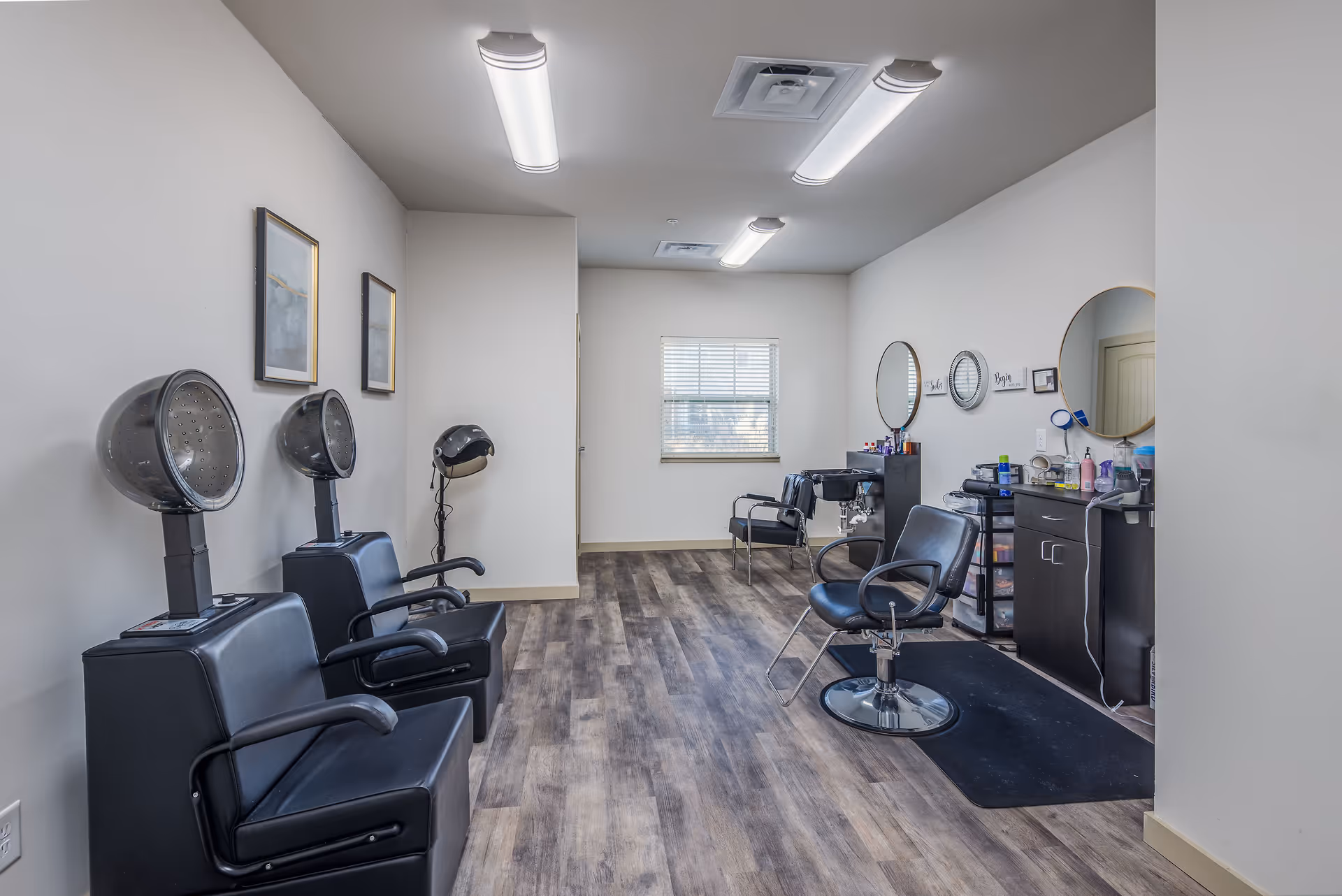 Interior salon room with hooded hair dryers, styling chairs, mirrors and workstations on wood-look flooring.