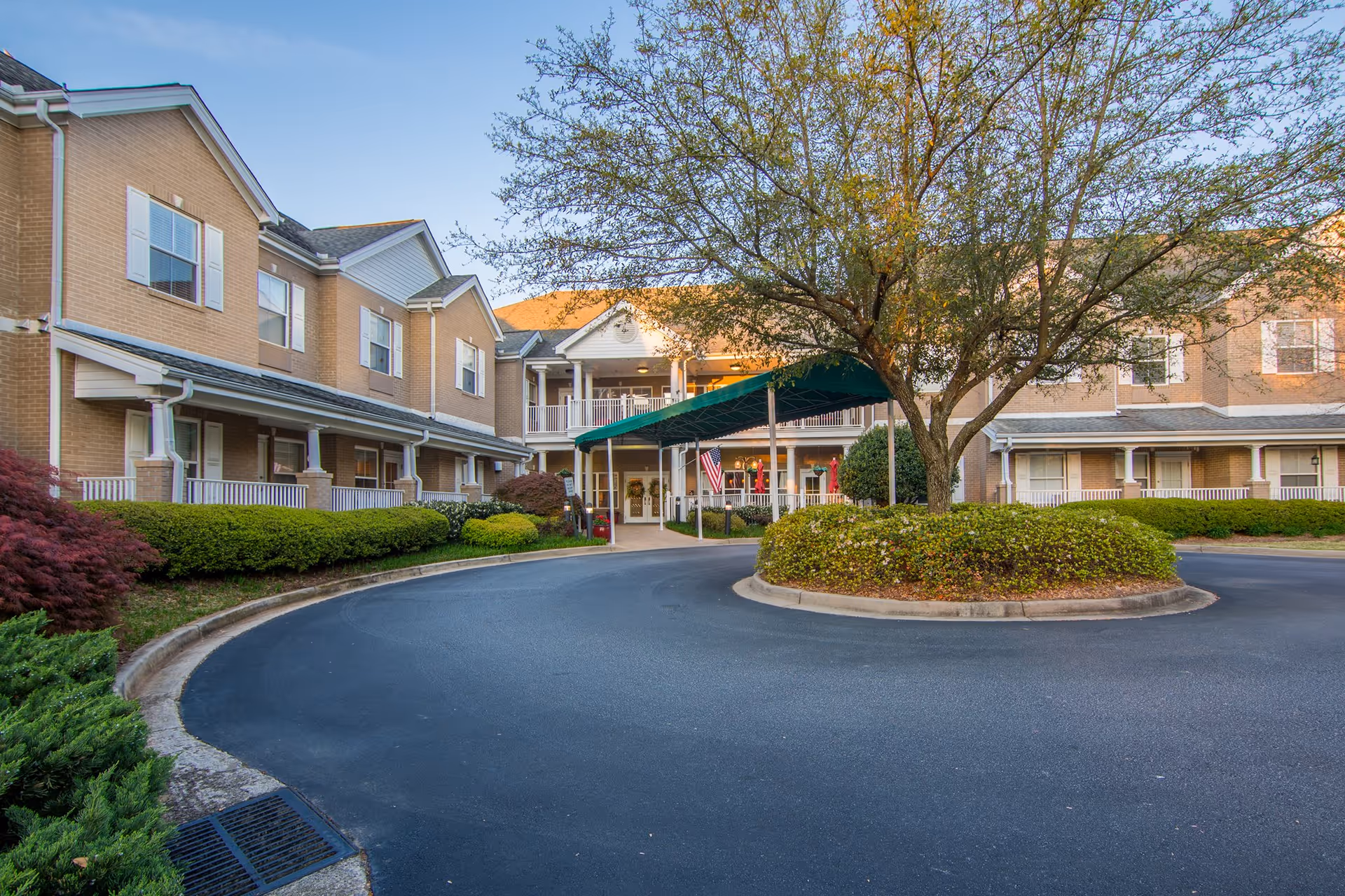 Exterior view of Senior Garden Assisted Living facility showing a two-story brick building with white trim and multiple windows. There is a circular driveway with a landscaped island featuring a tree and bushes in front of the main entrance, which has a green canopy. The sky is clear and blue.