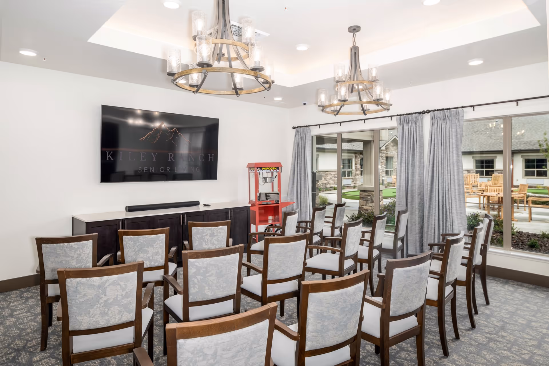 A senior living facility meeting room with rows of wooden chairs upholstered in light fabric facing a large flat-screen TV mounted on a white wall. The TV displays the logo and name 'Kiley Ranch Senior Living.' There are two large chandeliers hanging from the ceiling, a red popcorn machine in the corner, and large windows with gray curtains showing an outdoor patio area with wooden tables and chairs.