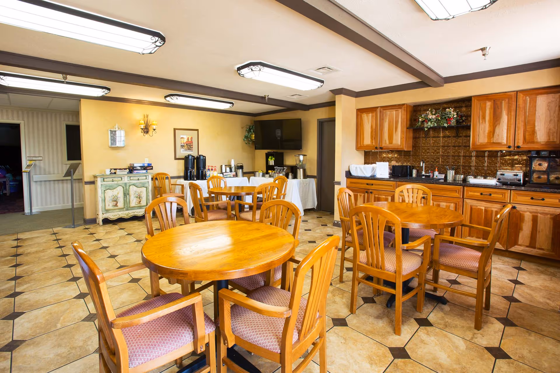 A dining area in a senior living facility with several wooden round tables and chairs with cushioned seats. The room features tiled flooring, wooden cabinets along one wall, and a counter with various kitchen appliances and a floral arrangement. There is a TV mounted on the wall and a side table with coffee dispensers and condiments.