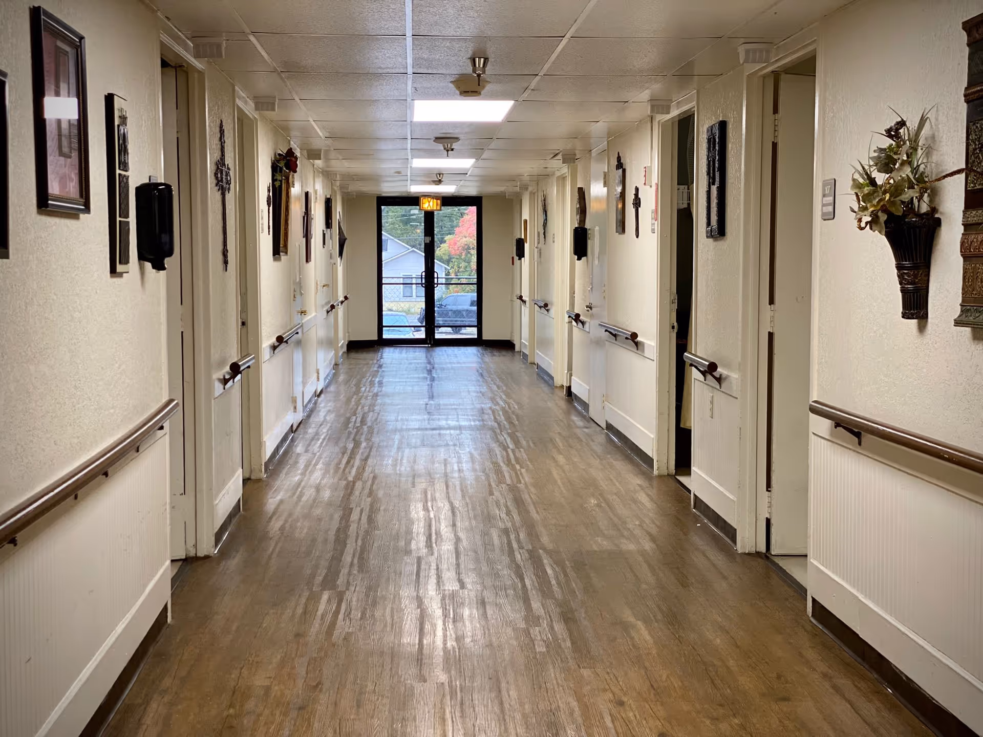 A long, well-lit hallway in a healthcare facility with wooden flooring and white walls. Doors line both sides of the corridor, each equipped with handrails. The walls are decorated with framed pictures, crosses, and wall-mounted flower arrangements. At the end of the hallway, there is a glass door with an exit sign above it, showing a glimpse of the outside.