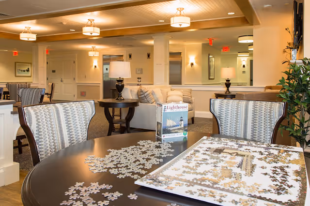 A cozy common area in a senior living facility with a round table in the foreground featuring a partially completed jigsaw puzzle and puzzle box. The background shows comfortable seating with sofas, armchairs, side tables with lamps, and warm lighting fixtures on the ceiling and walls.