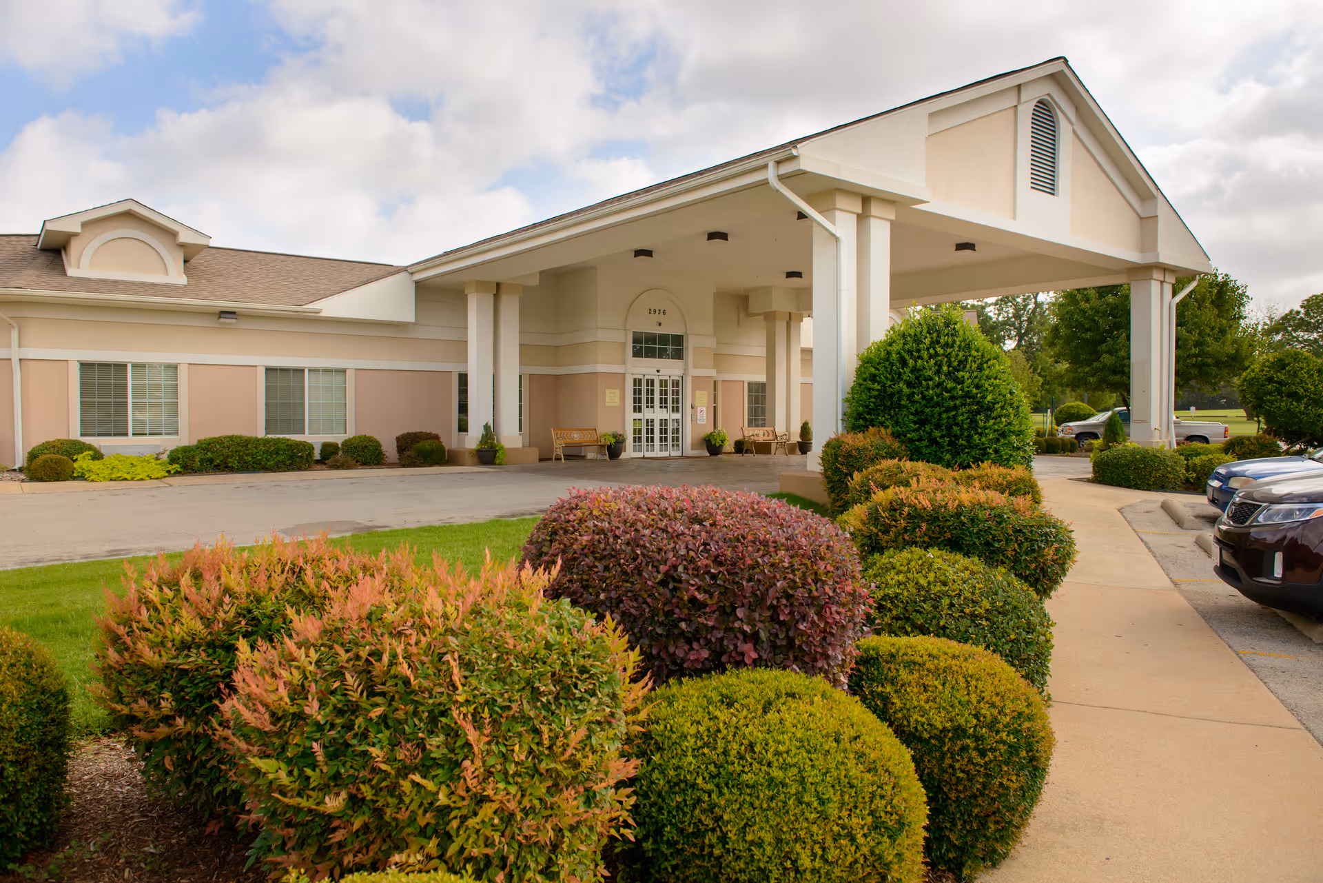 Front entrance of a single-story care facility with a covered portico, manicured shrubs, benches, and parked cars.