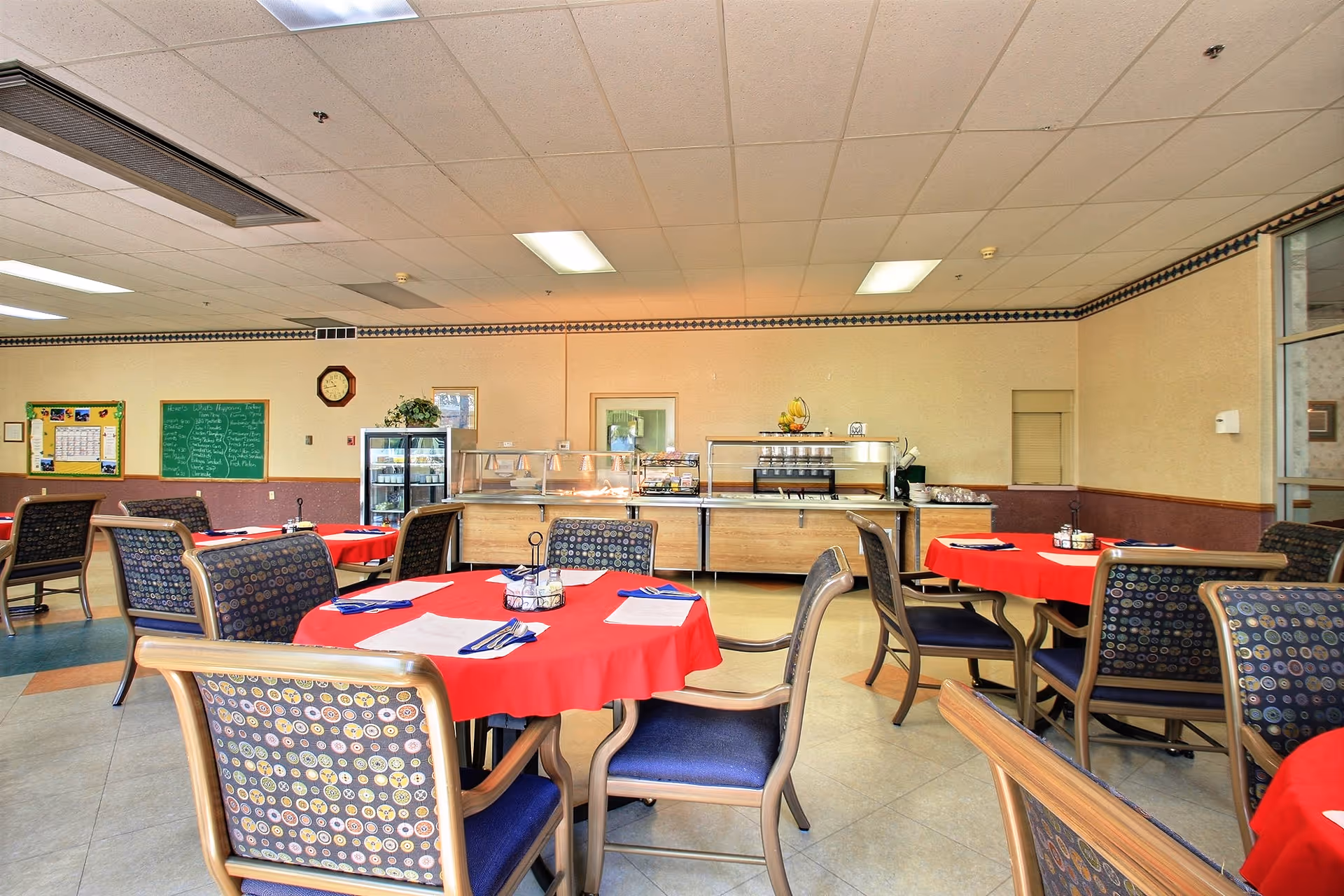 Dining area with round tables covered in red tablecloths, patterned chairs, and a buffet serving counter along the back wall.