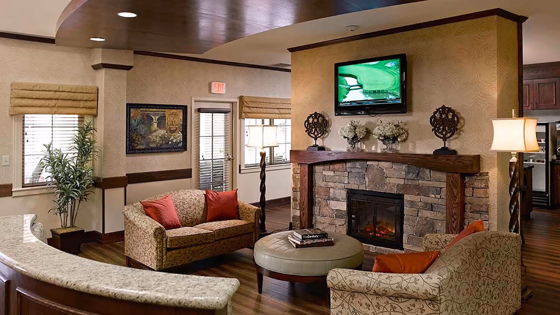 Cozy living room area with patterned sofas adorned with red and orange cushions, a round ottoman with books on top, a stone fireplace with a wooden mantle, and a flat-screen TV mounted above it. The room features warm lighting, a tall floor lamp, a potted plant, framed artwork on the wall, and windows with beige Roman shades.