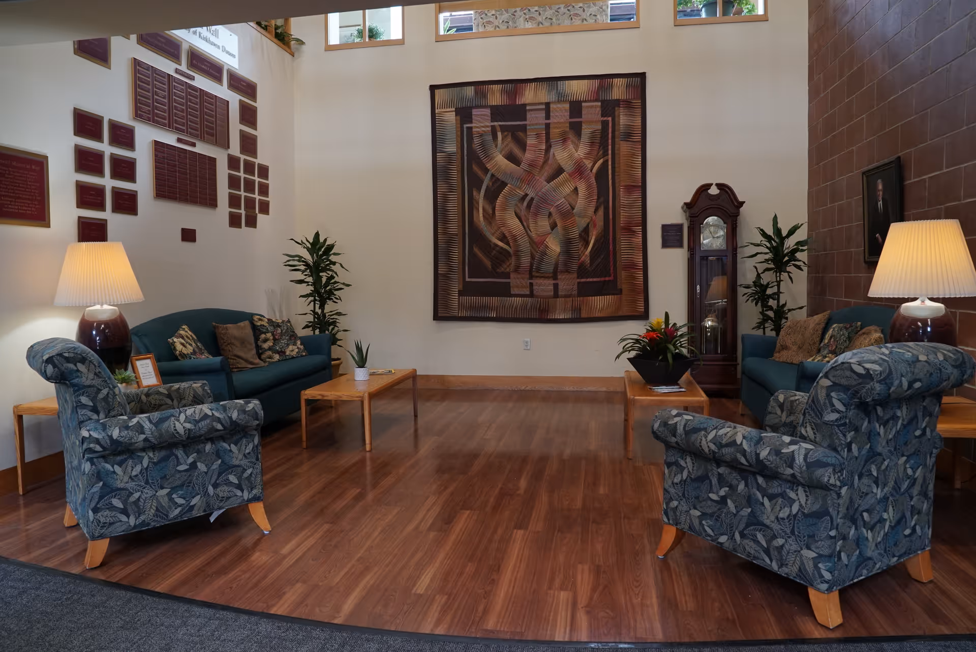 A cozy sitting area with two patterned armchairs and two green sofas arranged around wooden coffee tables. The room has wooden flooring, two table lamps with beige shades, potted plants, a large abstract wall hanging, and a tall grandfather clock. Plaques are mounted on the wall to the left, and a portrait hangs on the right wall.