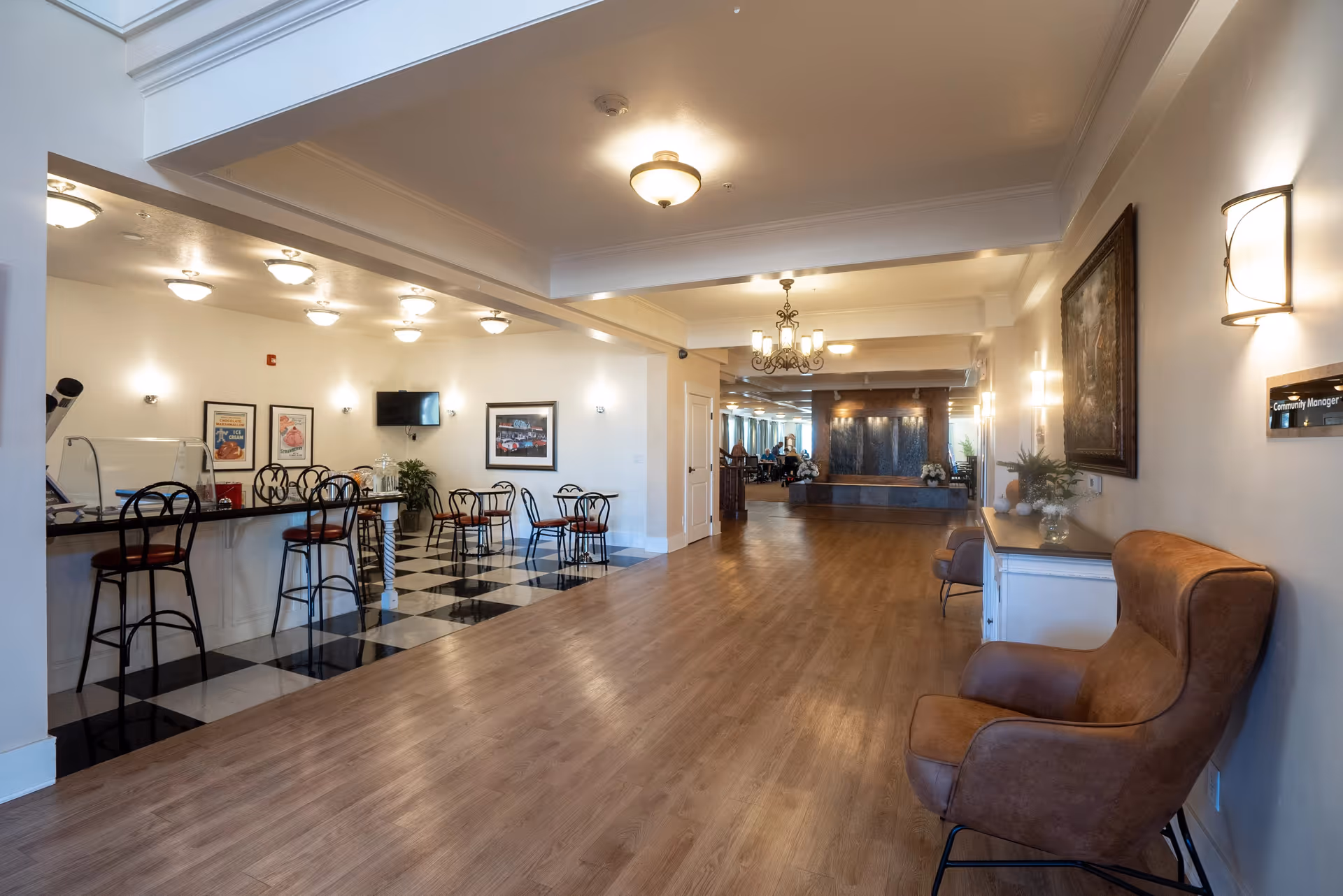 Interior view of a senior living facility showing a spacious hallway with wooden flooring and a seating area with brown armchairs on the right. On the left, there is a small dining area with black and red chairs and tables on a black and white checkered floor. The hallway leads to a common area with people seated in the background and a decorative water feature on the far wall. The walls are adorned with framed pictures and light fixtures.