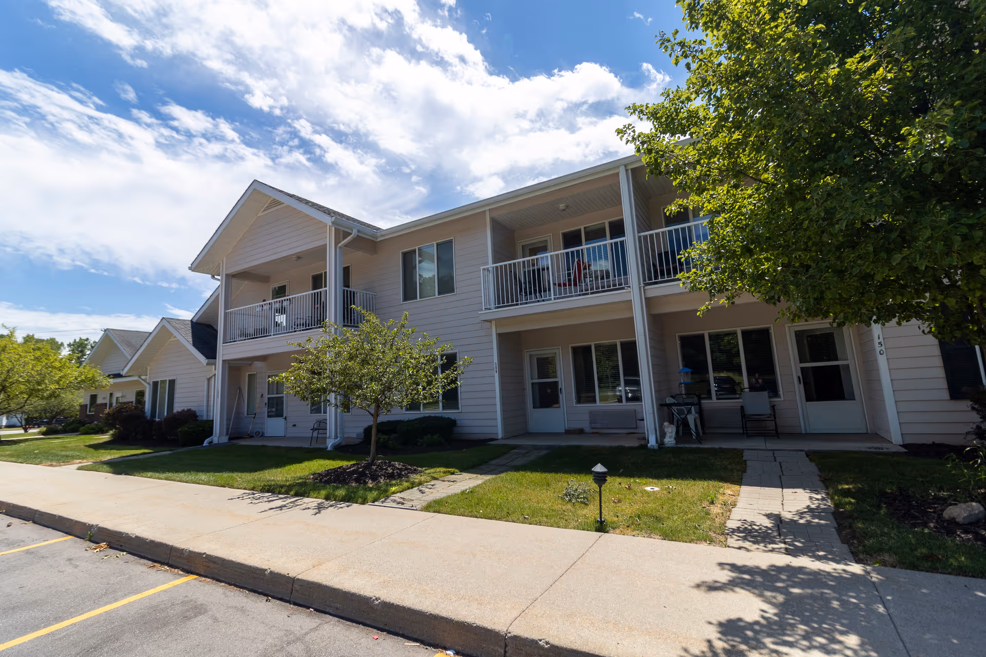 Front exterior of a two-story senior living building with upper balconies, ground-level patios, trees, and a sidewalk.