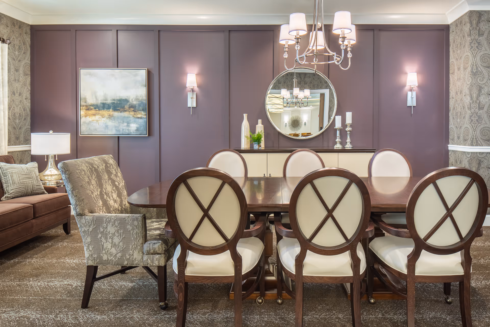 A dining room with a large wooden table surrounded by six chairs with white cushioned backs and seats. The room features a purple accent wall with a round mirror and two wall sconces, a sideboard with decorative vases and candles, a patterned armchair, a brown sofa with cushions, a table lamp, and a framed abstract painting.