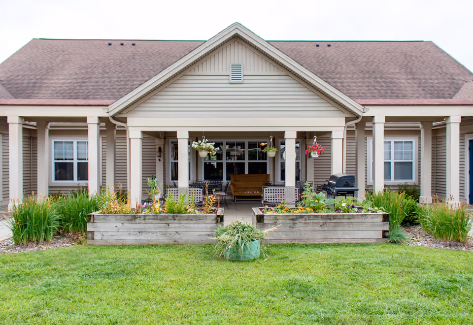 Front exterior of a single-story senior living building with a covered porch, hanging baskets, seating and raised flower beds.