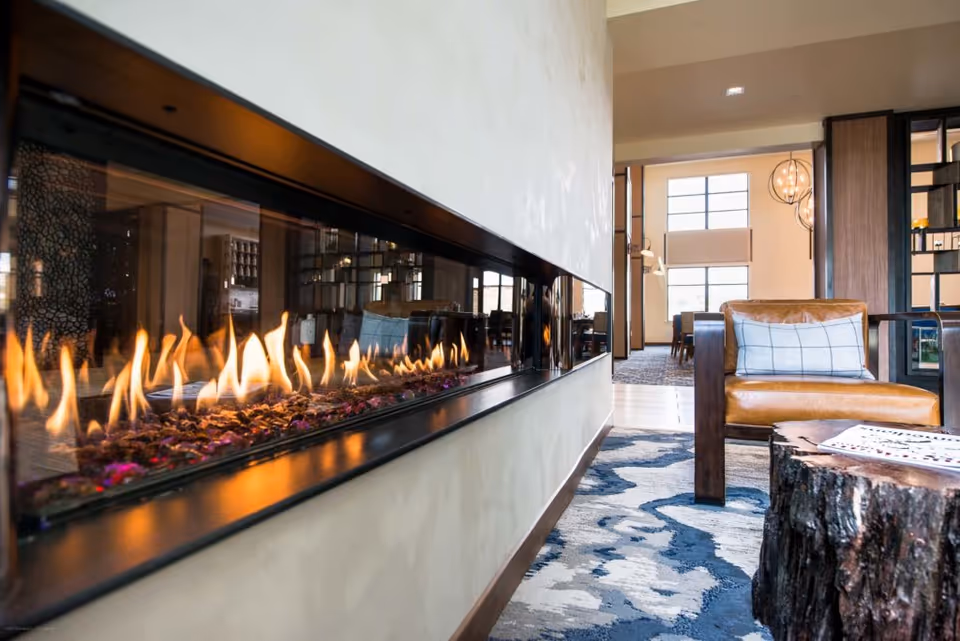 Modern indoor living area featuring a long, horizontal gas fireplace built into a wall. In the foreground, there is a leather armchair with a plaid cushion and a rustic wooden coffee table on a patterned blue and white rug. The background shows large windows and additional seating in a well-lit, spacious room.