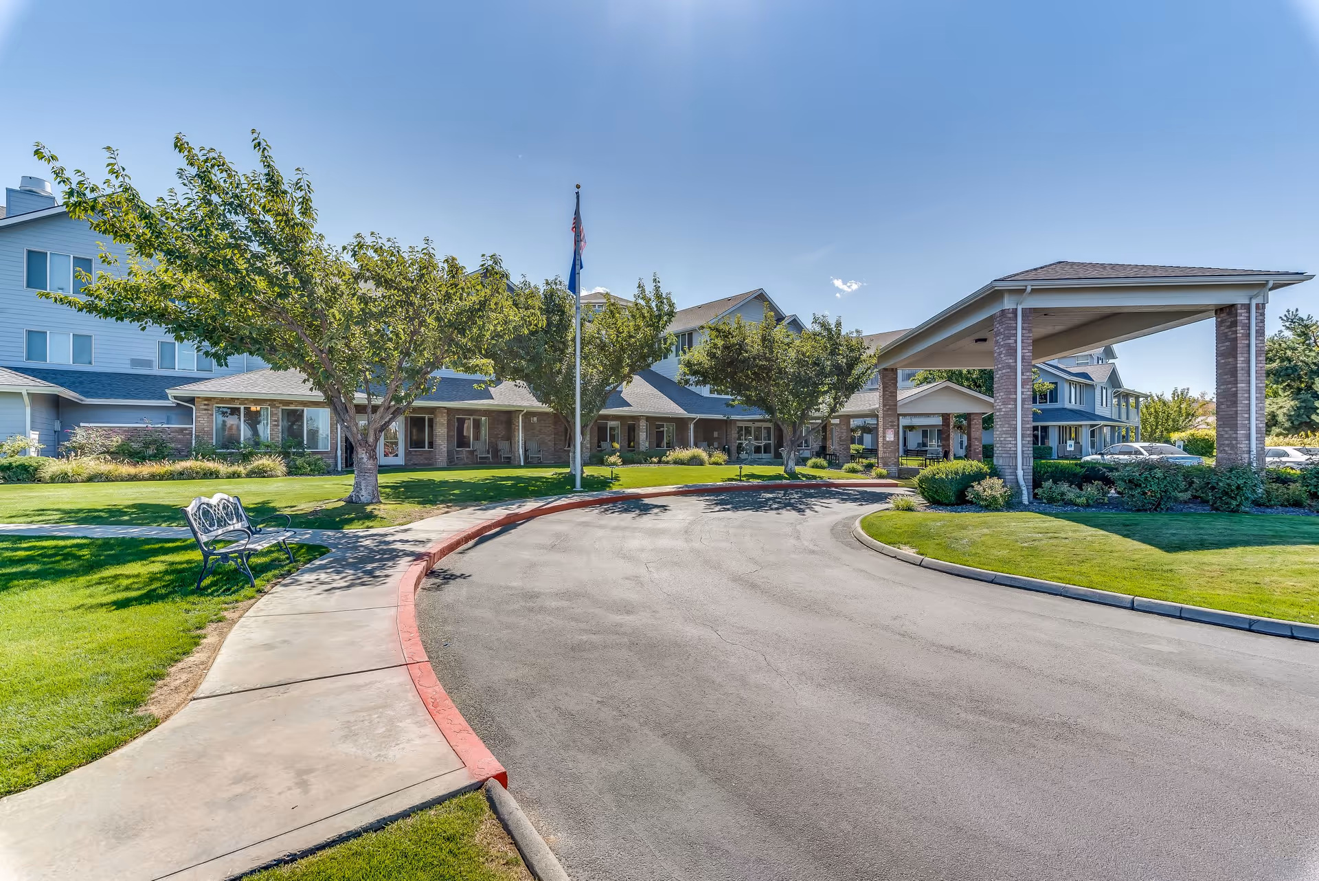 Exterior view of Solstice Senior Living at Kennewick showing a curved driveway with a covered entrance, green lawns, trees, and a bench under a clear blue sky.