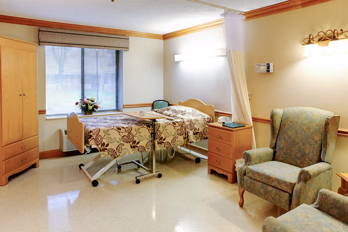 A well-lit patient room in a healthcare facility featuring a hospital bed with floral bedding, a bedside table with a phone and a book, a floral upholstered armchair, a wooden wardrobe, and a window with blinds and a flower arrangement on the windowsill.