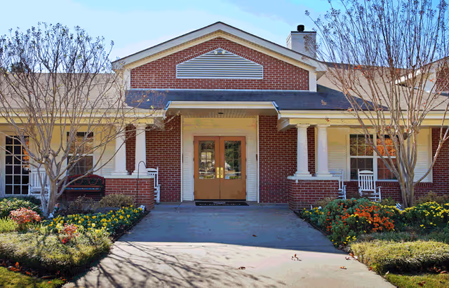 Front exterior view of a single-story brick building with a covered entrance supported by white columns. There are two leafless trees and flower beds with colorful flowers on either side of the concrete walkway leading to double wooden doors. The sky is clear and blue.