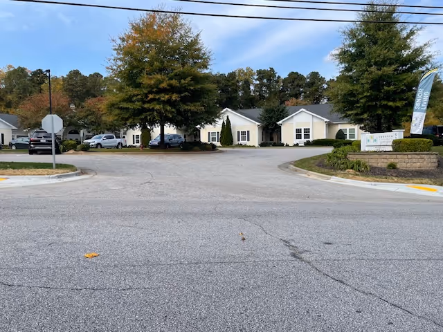 View of the entrance area to Cadence North Raleigh senior living facility, showing a paved driveway with parked cars, trees with autumn foliage, and single-story light-colored buildings in the background. A sign and a flag indicating 'Senior Living' are visible on the right side near a landscaped area.