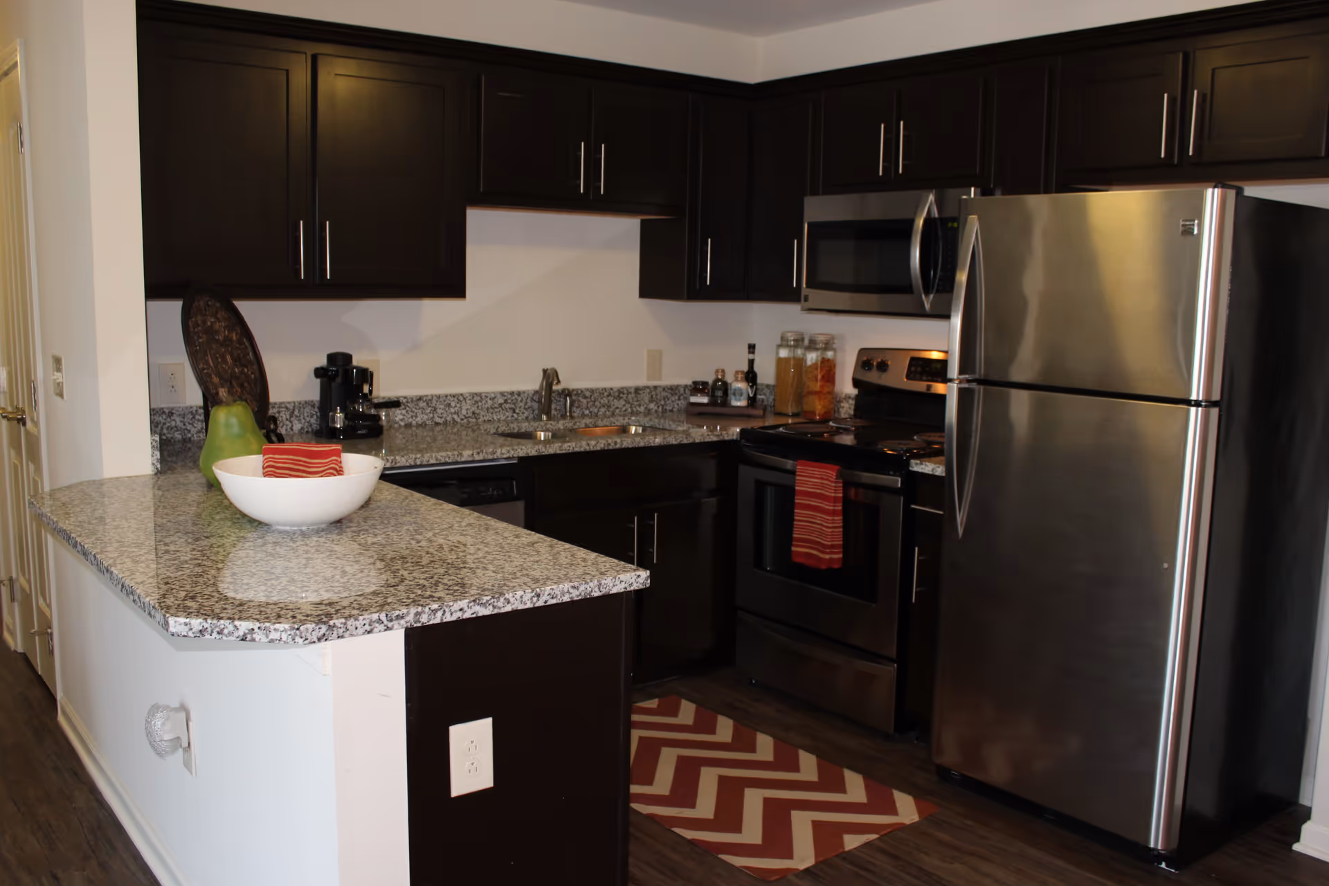 A modern kitchen featuring dark cabinets, stainless steel appliances, and a granite countertop with a bowl of fruit and decorative items.