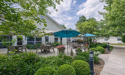 Outdoor patio area at Arden Courts - ProMedica Memory Care Community (Westlake) featuring tables with umbrellas, chairs, lush green bushes, trees, and a paved walkway under a partly cloudy sky.