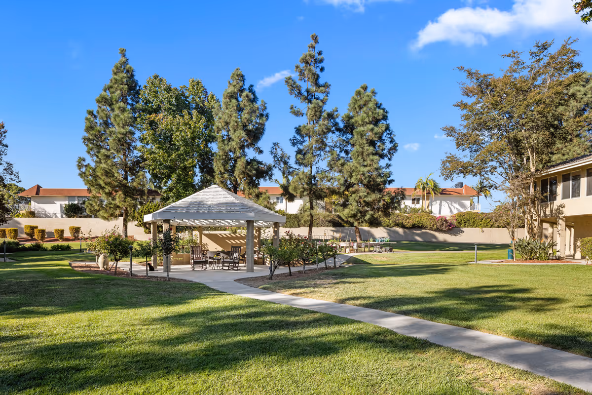 Outdoor area of a senior living facility with a white gazebo surrounded by trees and green grass. There are chairs under the gazebo and a paved walkway leading to it. The sky is clear and blue.