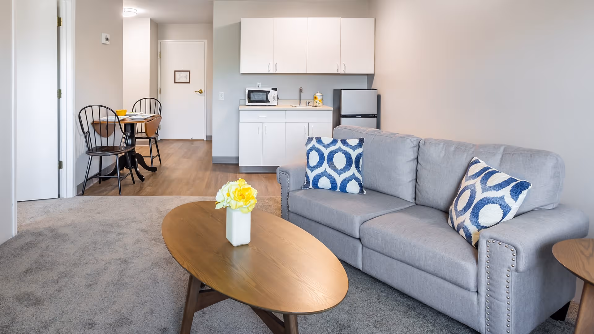 A cozy living area in a senior living facility featuring a gray couch with two blue and white patterned pillows, a wooden oval coffee table with a small vase of yellow flowers, a kitchenette with white cabinets, a microwave, and a mini fridge, and a small dining area with a round table and two chairs in the background.