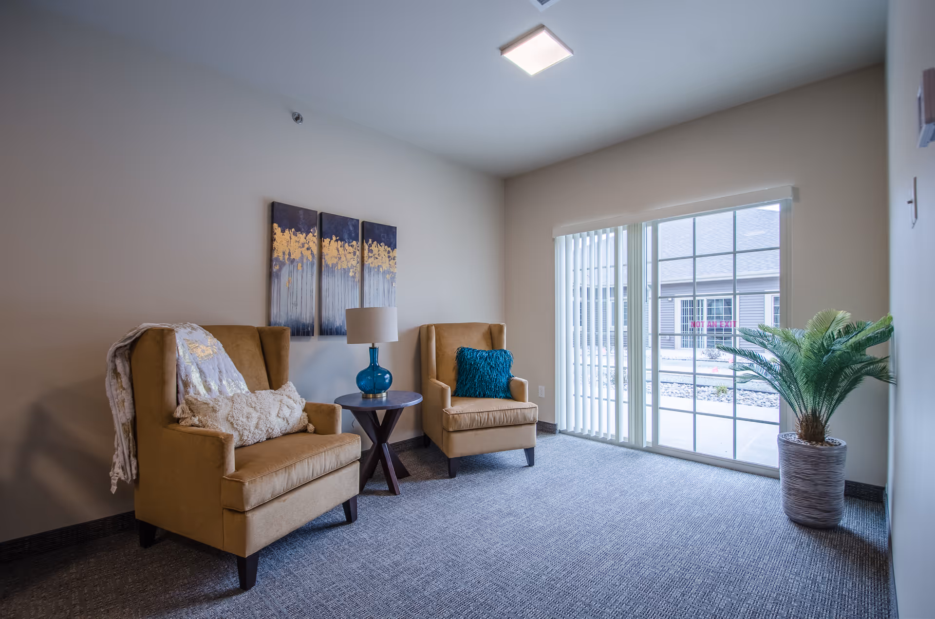 A cozy sitting area with two tan armchairs, one with a white throw blanket and pillow, the other with a blue decorative pillow. Between the chairs is a small round wooden table with a blue glass lamp. On the wall above the chairs is a three-panel abstract painting with gold and blue tones. To the right, there is a large window with vertical blinds and a potted plant on the floor. The room has light-colored walls and carpeted flooring.