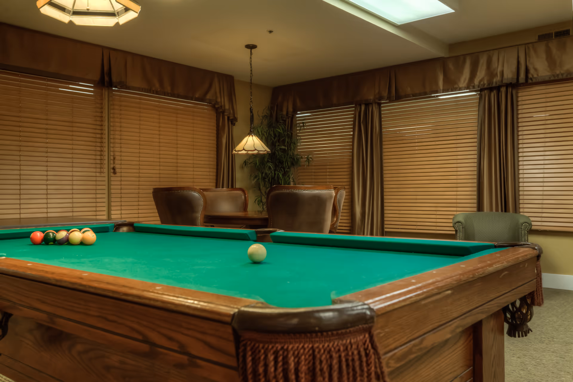 A room with a green felt pool table in the foreground, a set of billiard balls arranged on the table, and a cue ball. In the background, there are brown leather chairs around a small table, large windows with closed wooden blinds and brown curtains, a hanging lamp, and a potted plant in the corner.